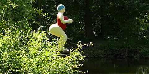 Am Rande des Teiches steht die lebensgroße Skulptur einer dicken Frau mit einem knallroten Badeanzug und einer Badekappe auf dem Kopf. Sie steht tief in den Knien und ist nach vorne geneigt, als wenn sie jeden Augenblick ins Wasser springen wollte.