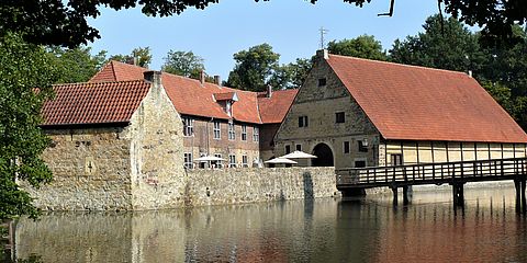 Im Bildvordergrund liegt der Burggraben. In der Bildmitte ist die Vorburg zu sehen. Rechts im Bild führt eine Holzbrücke von der Vorburg über die Gräfte in Richtung der Hauptburg, die selbst nicht zu sehen ist. 