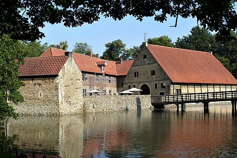 Im Bildvordergrund liegt der Burggraben. In der Bildmitte ist die Vorburg zu sehen. Rechts im Bild führt eine Holzbrücke von der Vorburg über die Gräfte in Richtung der Hauptburg, die selbst nicht zu sehen ist. 