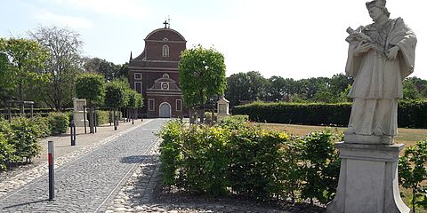 Langer gepflasterter Weg zur Kirche. Am Anfang des Weges steht eine Statur von dem Heiligen  Franziskus