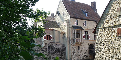 Der Blick geht über die Innengräfte auf die Hauptburg zu. Markant sind die dunklen Raseneisensteine im unteren Teil des Mauerwerks, der nach vorne gerichtete Erker und die münsterländischen Fensterläden in den Farben der Familie Droste Vischering rot und weiß.