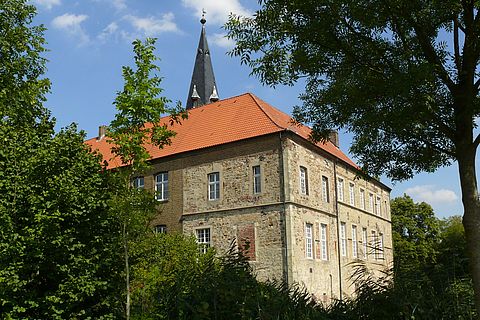 Hinter den Bäumen des Burggrabens erscheint die helle und sehr hohe Wand des Südflügels. Dahinter hebt sich die Turmspitze gegen den blauen Himmel ab.