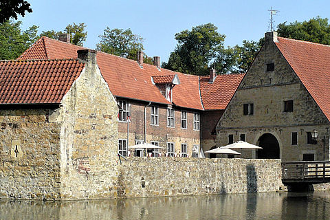 Im Bildvordergrund liegt der Burggraben. In der Bildmitte ist die Vorburg zu sehen. Rechts im Bild führt eine Holzbrücke von der Vorburg über die Gräfte in Richtung der Hauptburg, die selbst nicht zu sehen ist. 