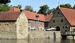 Im Bildvordergrund liegt der Burggraben. In der Bildmitte ist die Vorburg zu sehen. Rechts im Bild führt eine Holzbrücke von der Vorburg über die Gräfte in Richtung der Hauptburg, die selbst nicht zu sehen ist. 