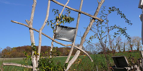 Holzgerüst aus Ästen, die einen Rahmen bilden. Darin ein Stück alte Plane mit einer handgeschriebenen Aufschrift zum Geissblatt.